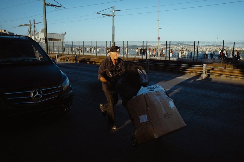 A man pushing a heavily loaded hand truck of cardboard boxes across a city bridge at sunset, passing a parked delivery van