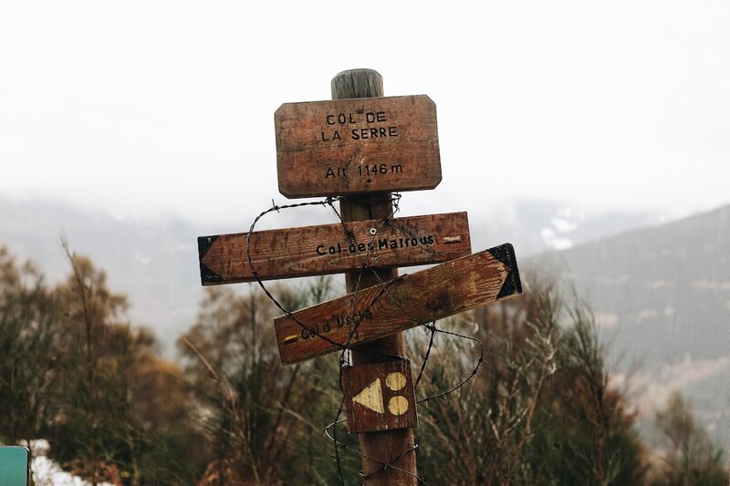 A weathered wooden signpost at a mountain pass in the rain, its arms lashed together with barbed wire