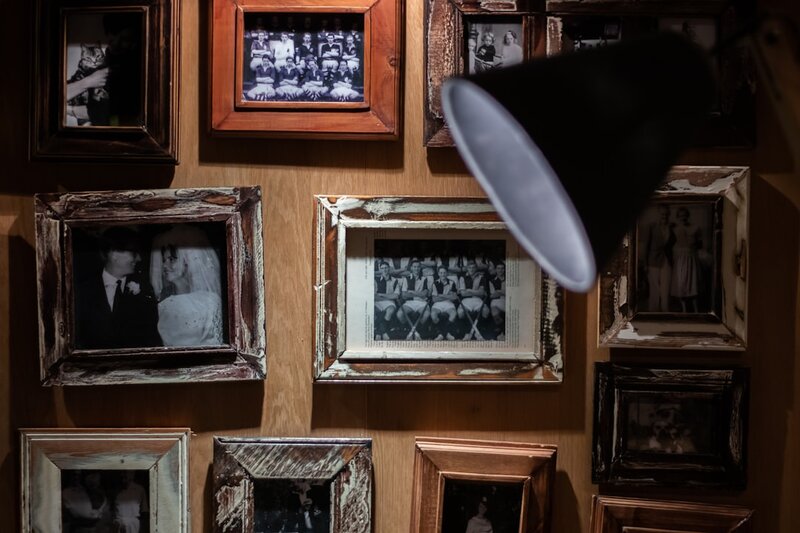 A wall of framed black and white photographs of people, lit by an angled desk lamp