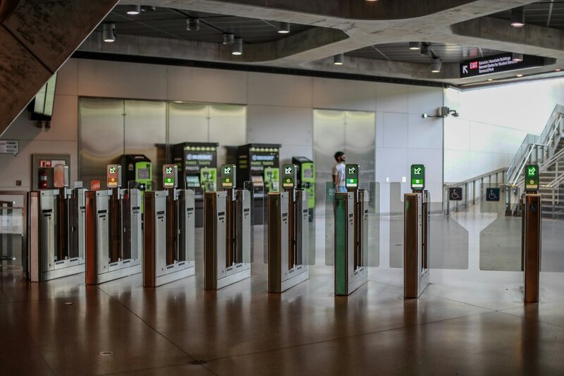 A row of closed subway fare gates in a station concourse, with a wide accessible gate standing open and unbarriered beside them