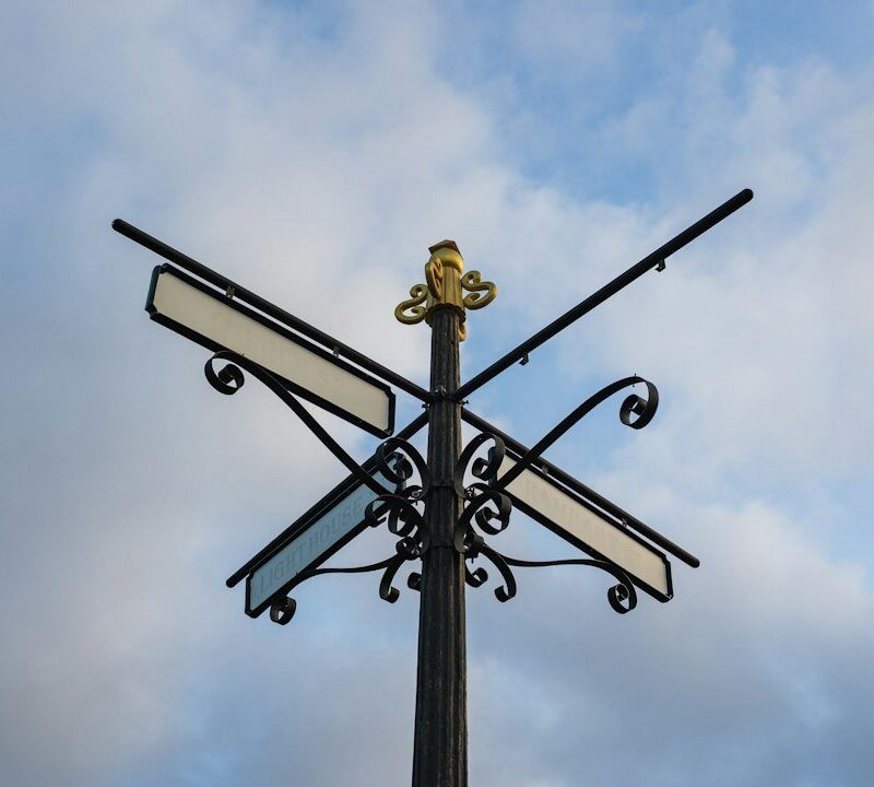 An ornate black four-way signpost photographed from below against a cloudy sky, its direction plates blank.