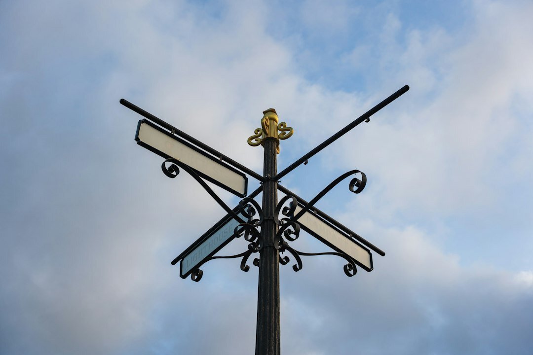 An ornate black four-way signpost photographed from below against a cloudy sky, its direction plates blank.