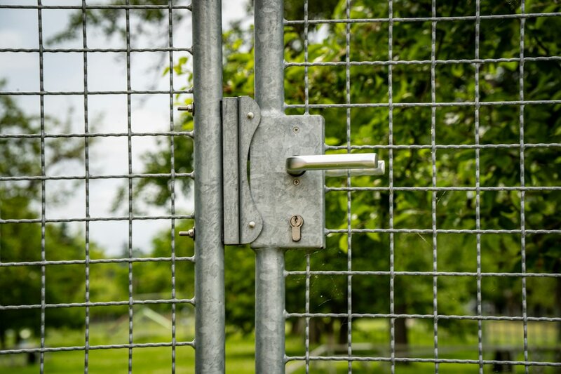 Close-up of a wire-mesh gate with a steel handle and lock mechanism, a green park behind it